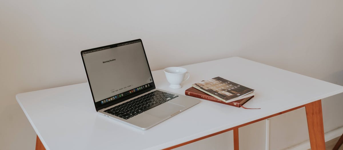 A minimalistic workspace featuring a laptop, coffee cup, and books on a white desk with wooden legs.